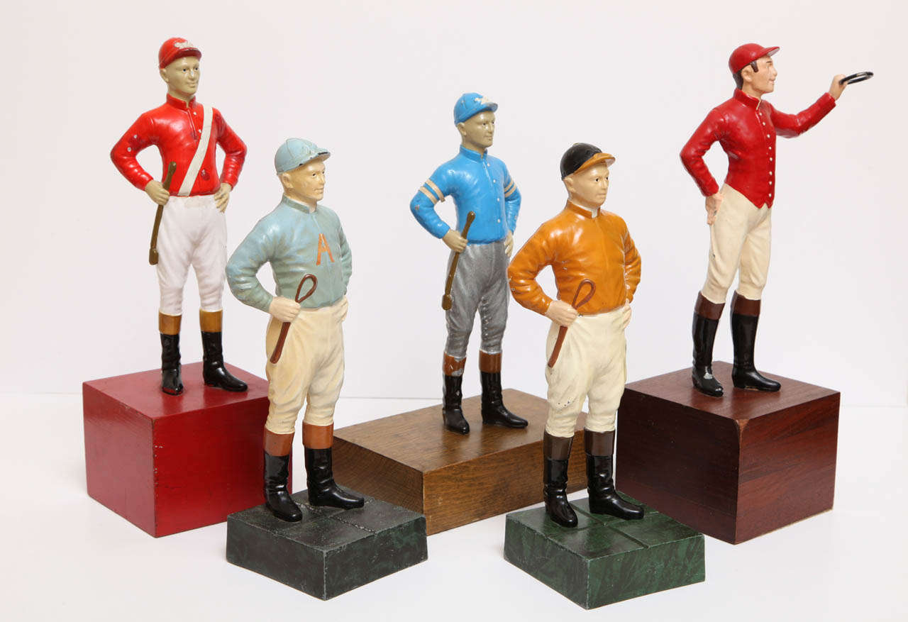 A group of iron lawn jockey sculptures in various colors. Each wearing their colorful signature silks. Four are holding a riding crop one has a ring in his hand for a lantern. Enamel is worn on a few of the jockeys but overall they are in excellent