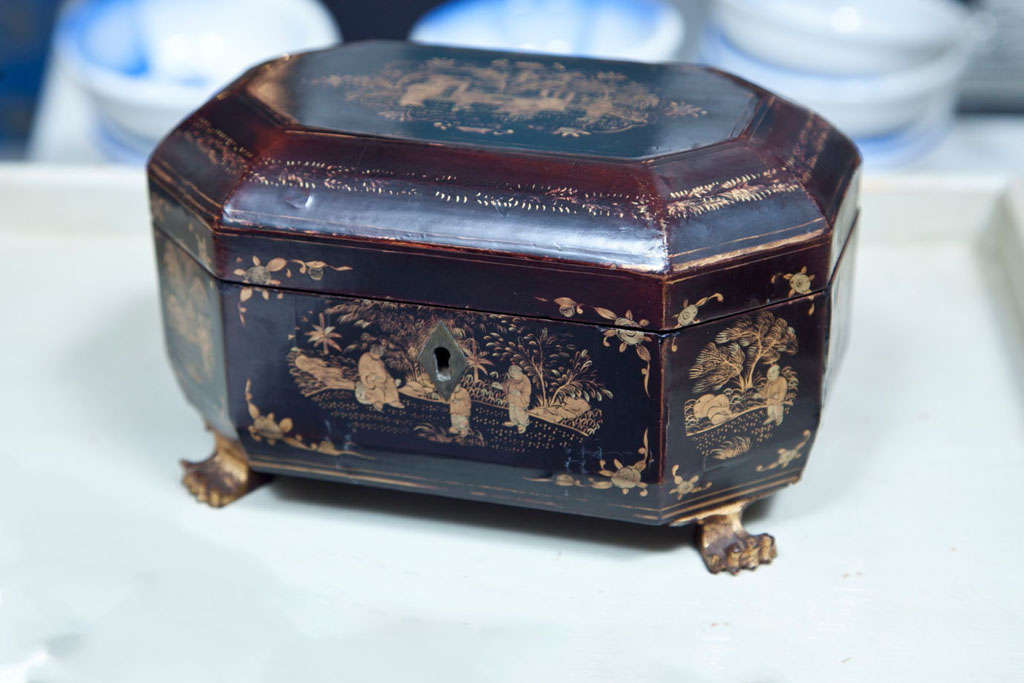 The burgundy / brown lacquer is decorated with gold depicting figures and buildings in a landscape. The caddy stands on four carved wooden feet in the shape of claws. The interior contains a single pewter canister insert with round raised lids.