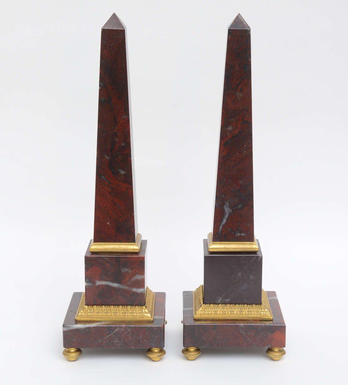 Large pair of obelisks made in a rosso antico marble with charcoal and white veining on brass platform footed square bases, with ferrule ball feet. Center of obelisks has acanthus leaf perimeter detail designs, with brass pencil surround and hobnail