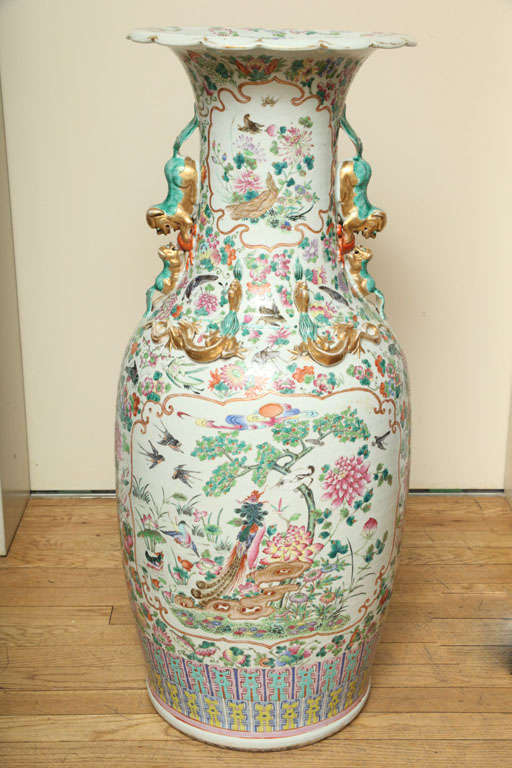 A pair of Chinese palace size porcelain vases, the flared lips with scalloped edges above tapered waist with foo dog handles. Vases decorated overall with traditional hand painted decoration in pink and green depicting trees, birds, and stylized