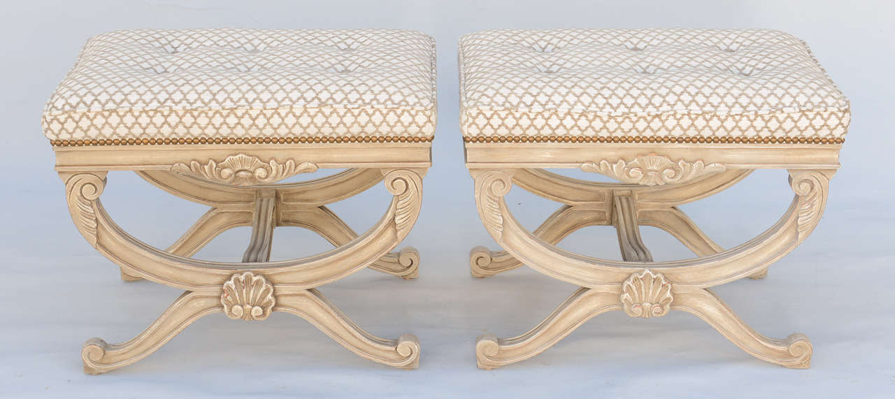 Pair of curule-form stools,or ottomans, each having rectangular boxed seats, upholstered with nailheads, on bases with painted finish; each fielded apron decorated by outcarved seashell, raised on foliate-headed reeded X-frame legs, joined by