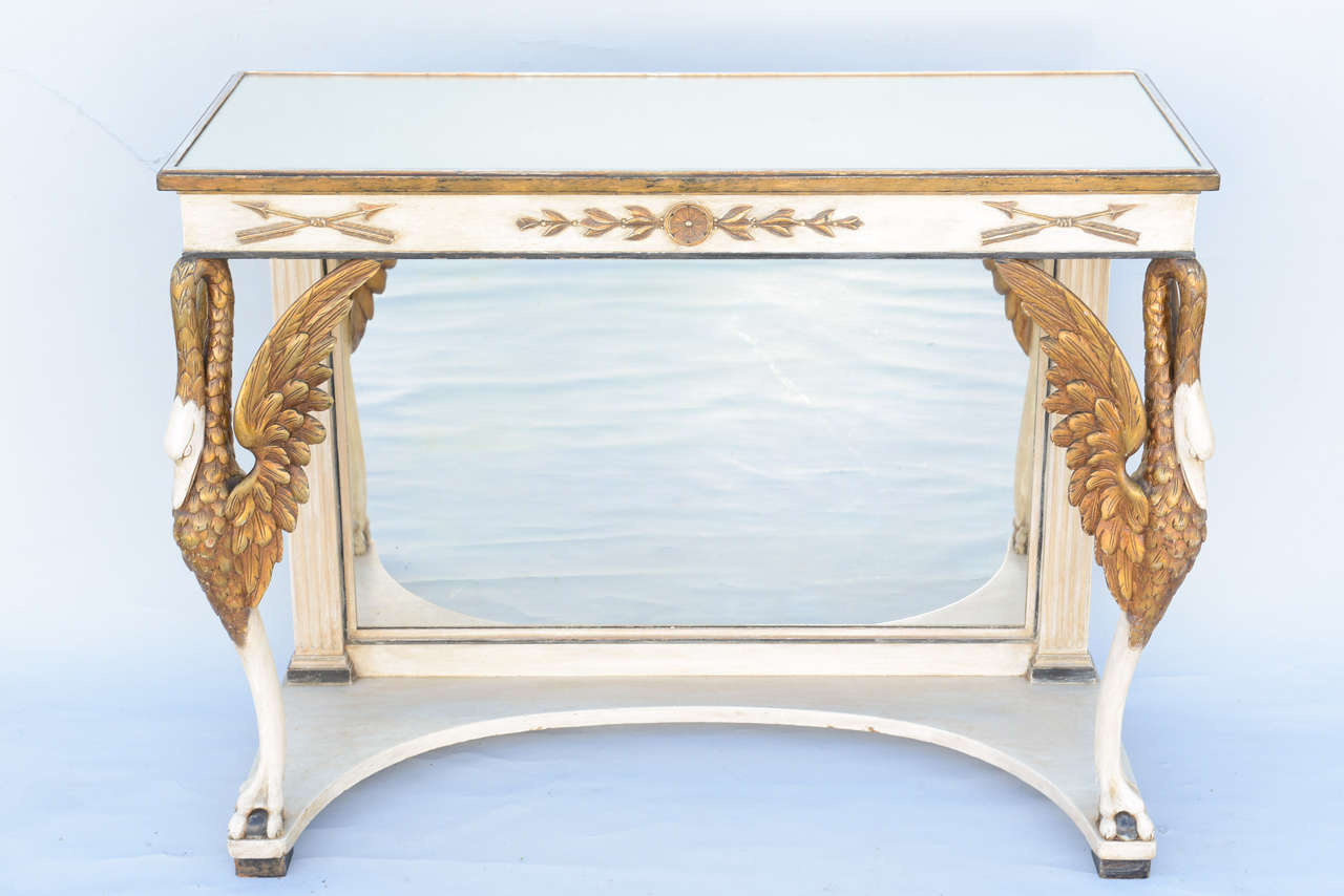 Console table, having a rectangular top of mirror, on painted and parcel gilt base, with mirrored back panel; its apron outcarved with arrows and husking, raised on elaborately carved swans with extended wings, ending in ball-and-claw foot, to its