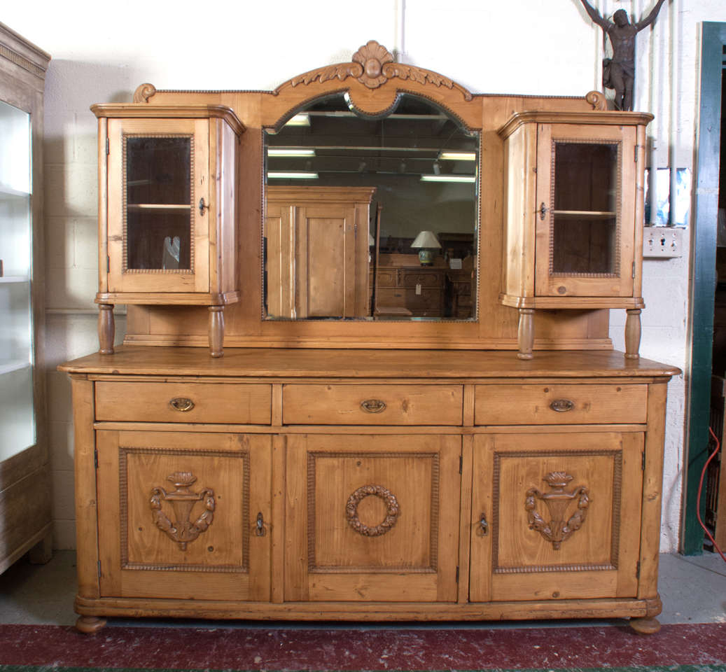 An unusual pine sideboard featuring a large central bevelled mirror flanked by two glazed display cupboards mounted on a base of three hand-cut dovetailed drawers above three panelled doors with with hand-carved appliques.