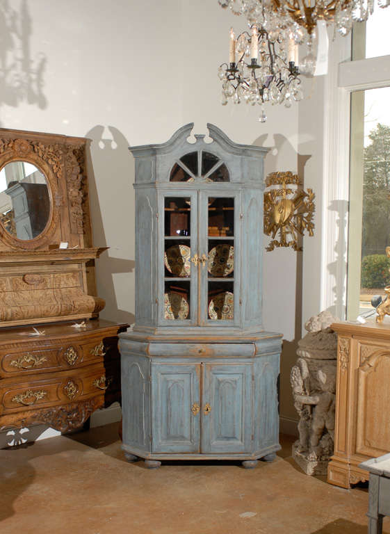 A Swedish, 18th century period baroque corner cupboard with broken pediment, arched window, canted corners and old glass. Painted in a light blue color with wood coming through, this Swedish corner cupboard features a tall upper cabinet, topped with