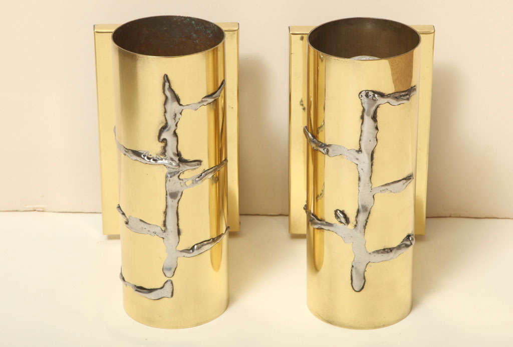 These whimsical yet modernist wall-mounted sconces are a fantastic highlight to any space. They feature soldered abstract silver forms on a polished brass cylinder. They cast a subtle amount of light that spills out from both the top and bottom.