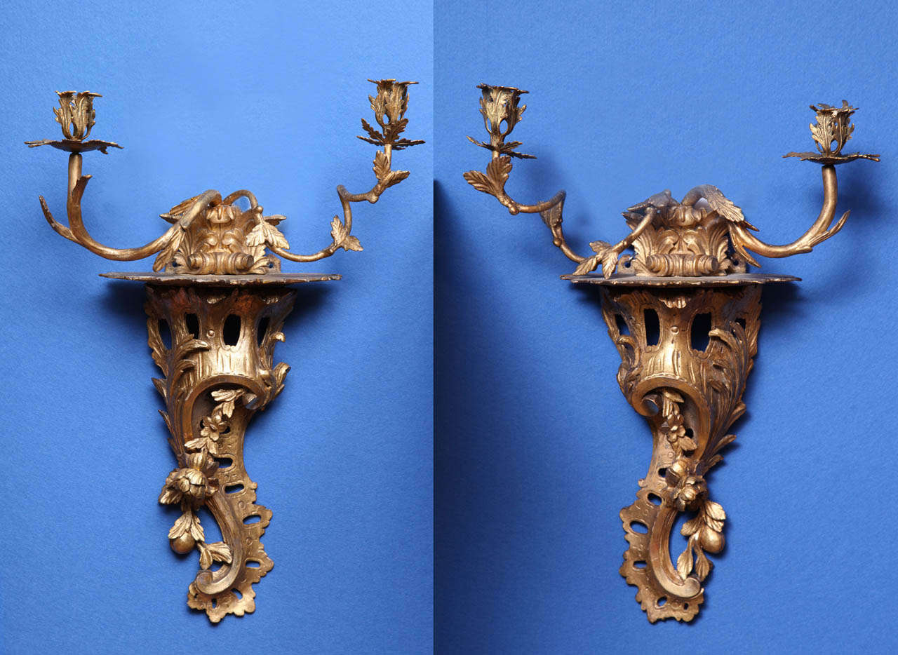 Rare Pair of Chippendale period carved Rococo giltwood wall lights, each having a pair of scrolling branch form arms with gilt metal cups and bobeches issuing from a central scroll, supported on a pierced rockwork bracket encrusted with 