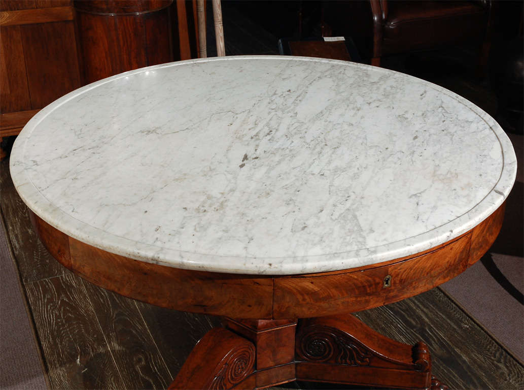 A gorgeous center table with a white and grey veined marble top on a round mahogany base with frieze drawers raised on an urn-shaped pedestal tripod base with scroll carvings and paw feet