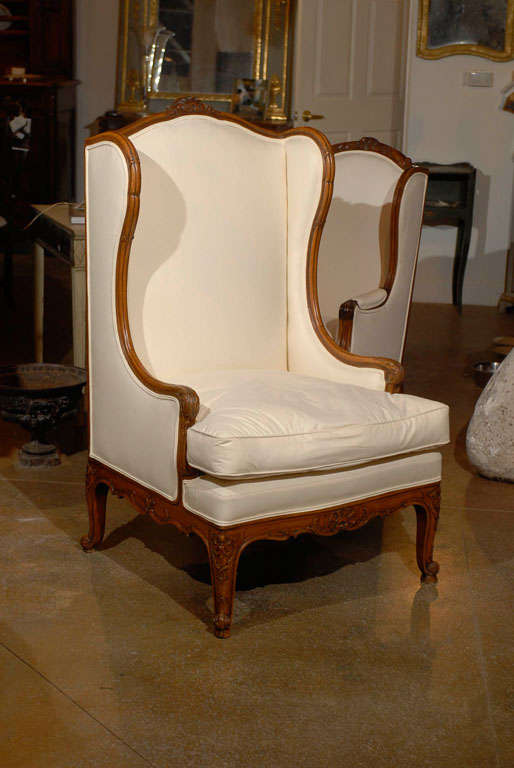 A French Louis XV style walnut wingback bergère chair from the 19th century with new upholstery. This French 'bergère à oreilles' features a straight back, adorned on the upper rail with a carved crest. The sides, designed to provide both privacy