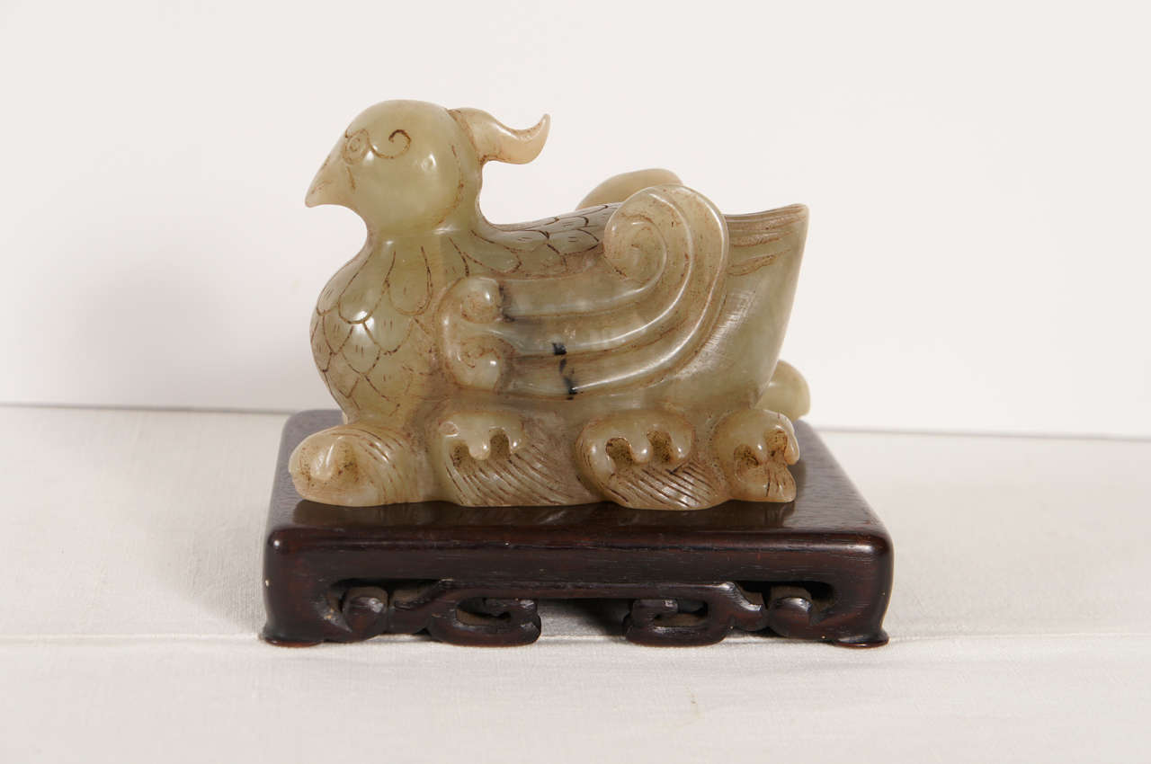 This nicely carved bird figure is done in an 18th-century style but is from the very late 19th century to early republic period. The stone is a soft mellow celadon green with dark brown inclusions. Carved on swirling waves the bird is alert and