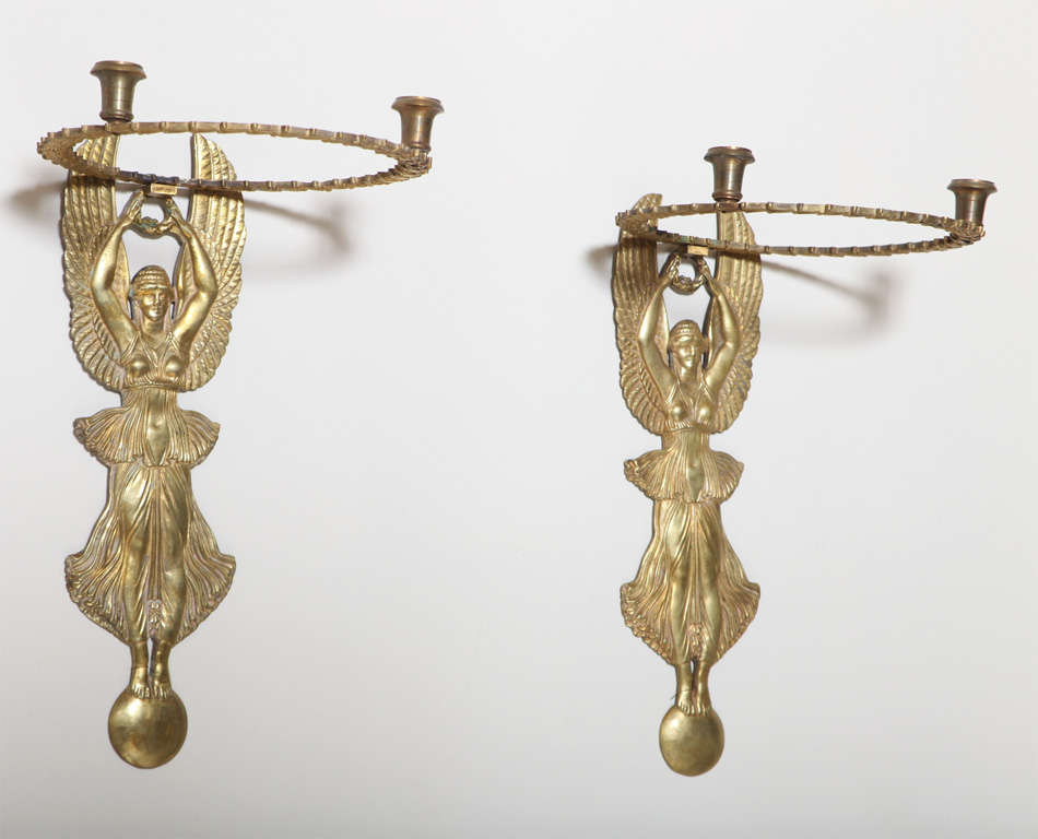 Pair of Empire style sconces, of bronze ormolu, each depicting a winged female figure with raised arms holding a wreath, joined to a chased ring with two candle pots.  Not electrified.