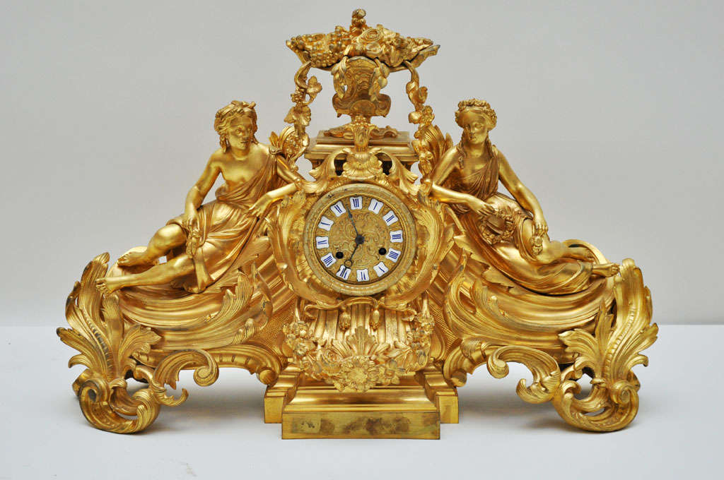 A Napoleon III Gilt Bronze 8 day French mantel clock, depicting two reclining classical Greco-Roman robed female figures with floral headdress, flanking a circular bronze dial face with enamel Roman numeral chapters, surmounted by a flowering urn.