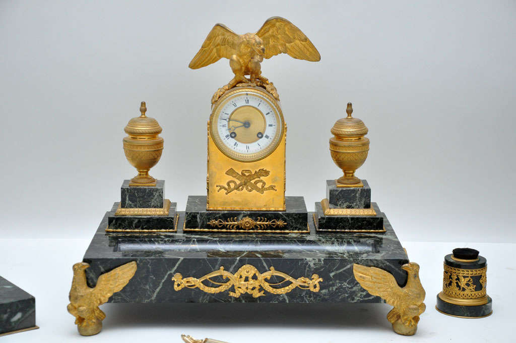 French Empire six-piece marble and gilded bronze desk set. Gilded bronze grand inkwell urns flank gilded bronze clock surmounted by a detailed eagle, adorned with smaller eagles that flank center holder for plume and letter opener; letter and file