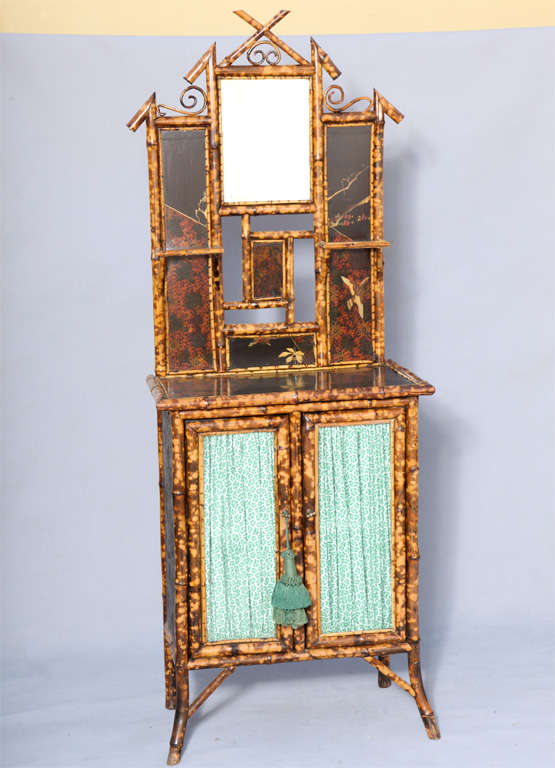 Cabinet, of burnt bamboo inset with japanned panels; its bevelled mirror surmounted by broken pediment and flanked by shelves, over cupboard base with double doors of glass, raised on splayed legs.