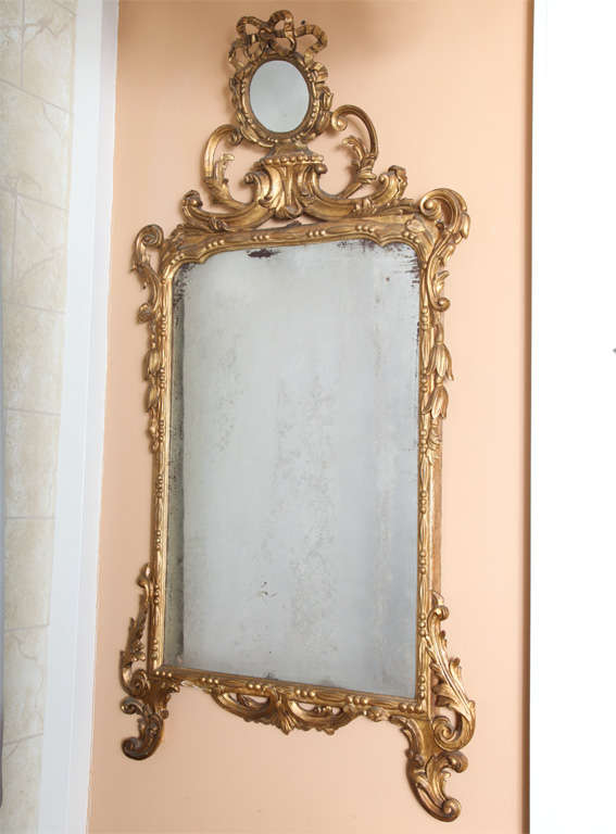 Mirror, of carved giltwood, having a rectangular bead-and-bar frame, surmounted by a pierced pediment of a round mirror plate and ribbons, supported by C-scrolls; husking and acanthus carved details decorate the frame.