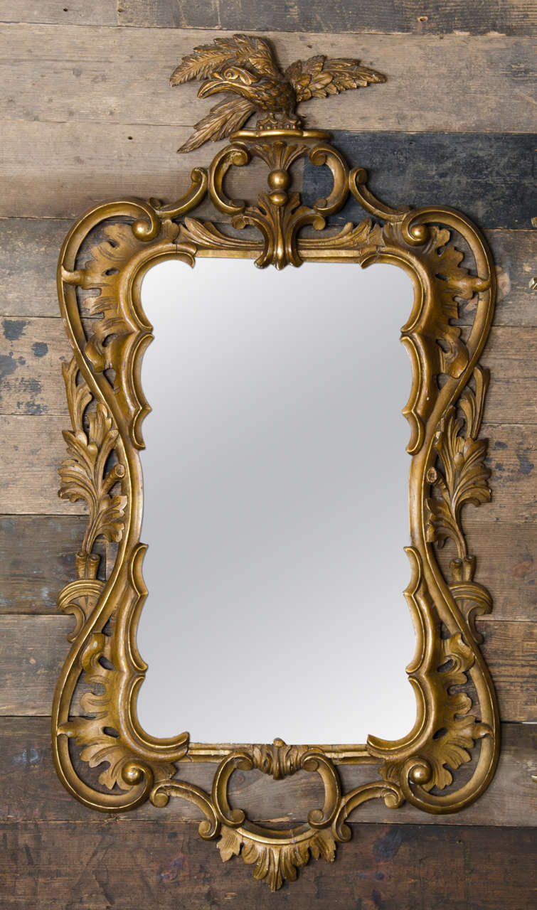 A decorative gilt framed mirror with a Rococo style. This gold framed mirror features ornate, scrolling decoration with an eagle perched at the top. The original antique glass has some characterful age spots but is in largely good condition.