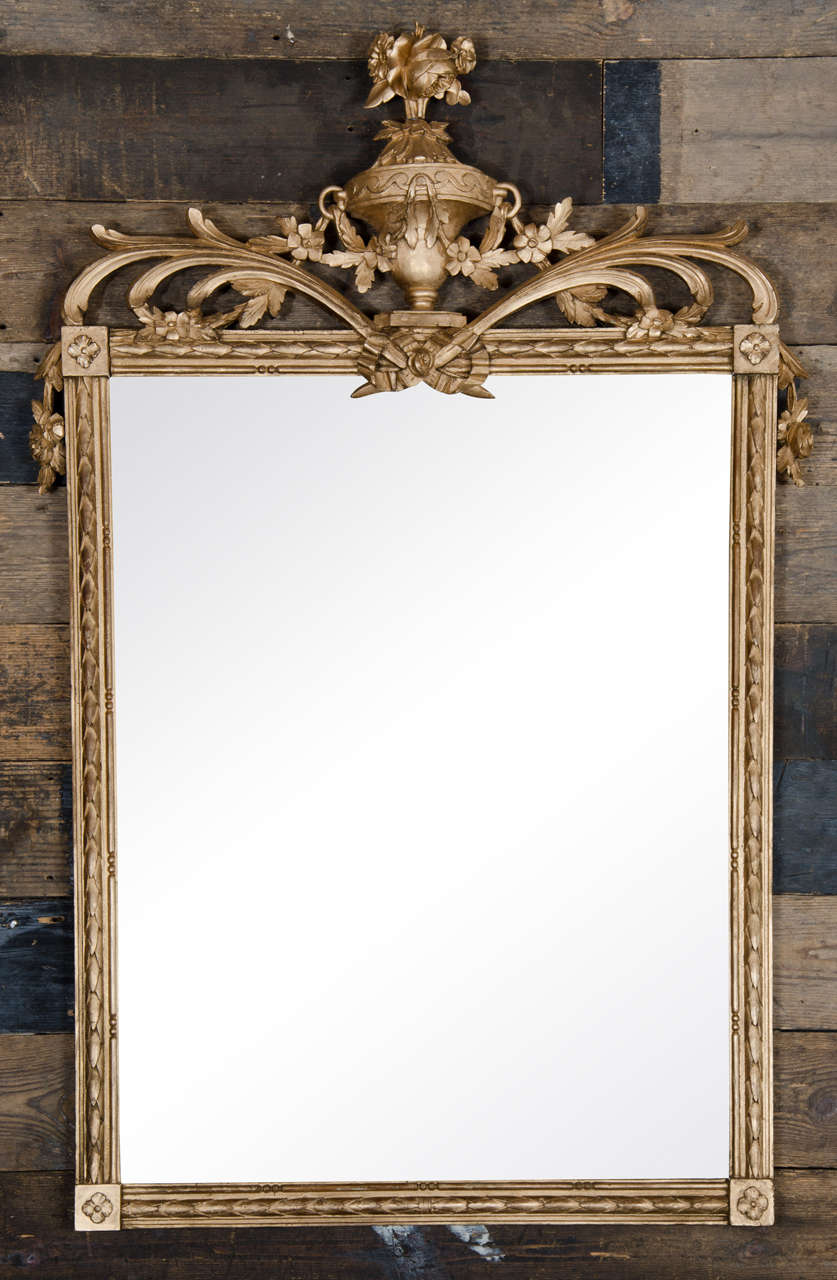 A beautifully decorative antique mirror with a gold gilt frame. This mirror has a rectangular frame with floral paterae at each corner and an elaborate floral decoration surrounding a central urn at the top.