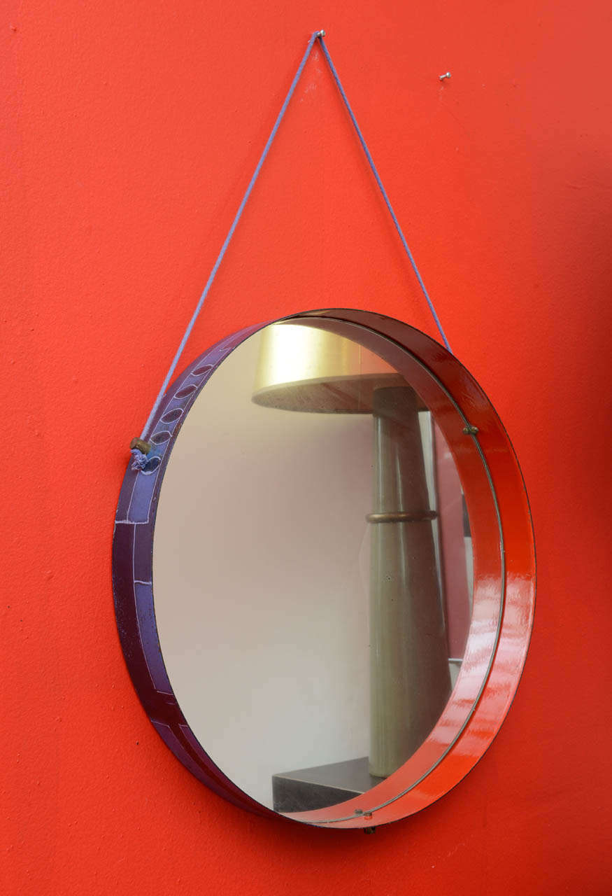 Round mirror decorated with abstract pattern on the outside and solid red interior band.
Hanging from a blue cord attached to the frame with brass fittings. Mirror is original and has some foxing.