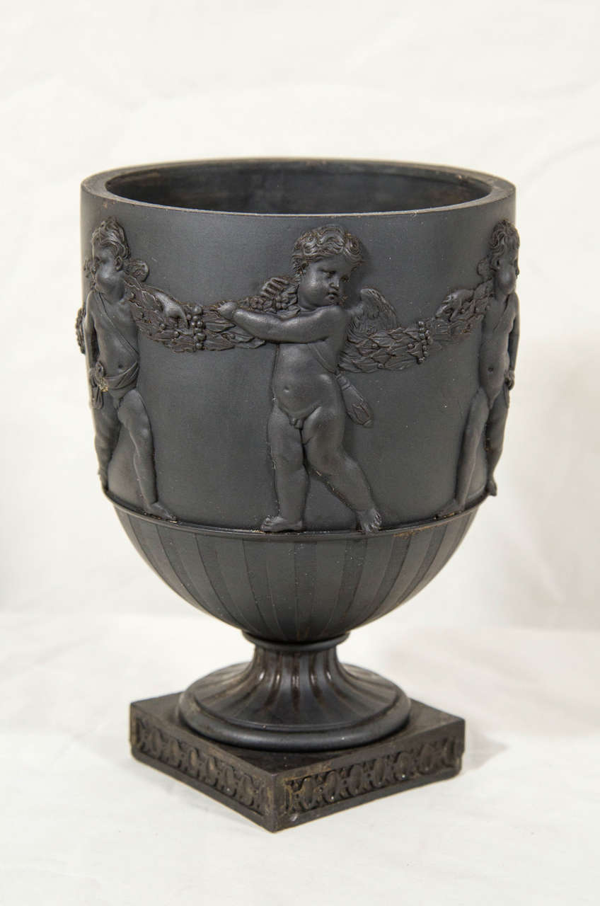 A Wedgwood & Bentley black basalt urn-shaped vase molded with decoration in strong relief showing angels suspending berried wreaths on their shoulders. They stand above a design of machine turned panels all raised from a square pedestal base. The