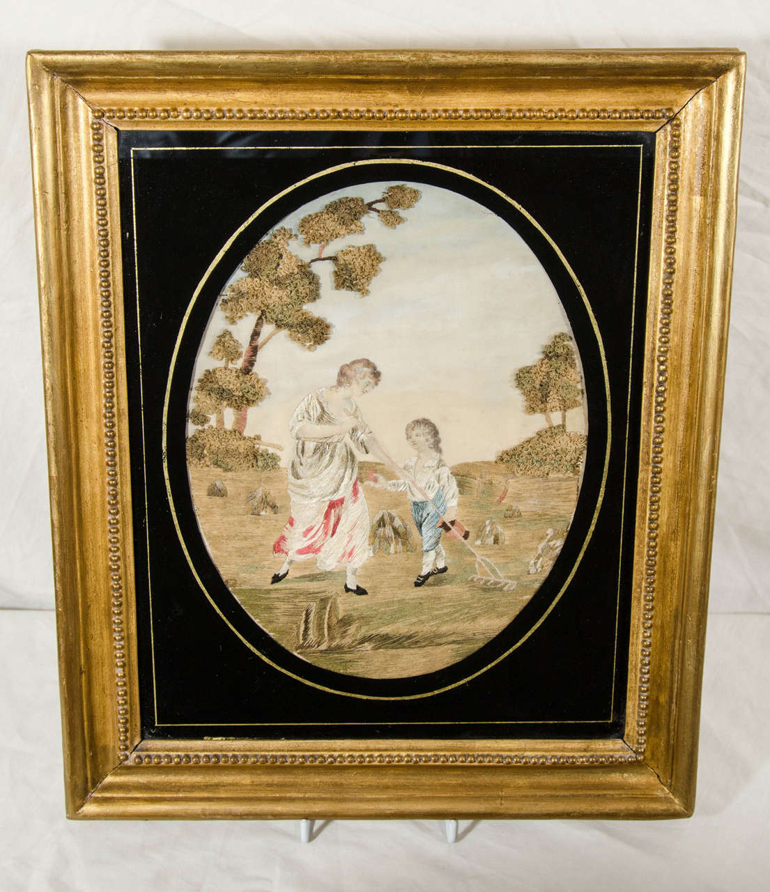 An English silk needlework embroidery in excellent condition and retaining good original colors. The needlework depicts a rural scene of a mother at work raking a field while her son brings her a bit of food and water. In one hand he carries a water