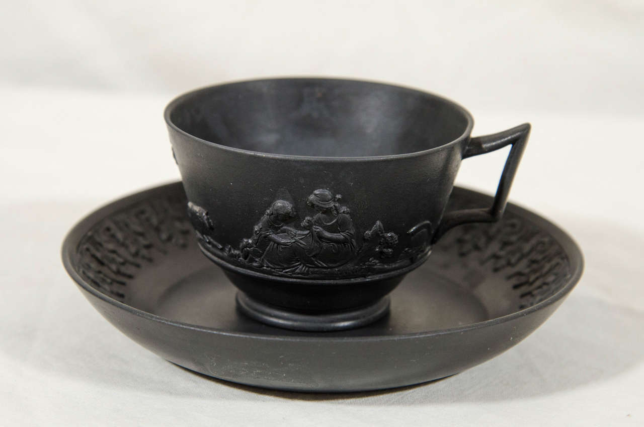 A dozen Wedgwood Black Basalt tea cups and saucers each cup with applied decoration showing a unique neoclassical scene with men and women in classical dress. The surfaces are lustrous and smooth, with a deep black sheen. The saucers impressed with
