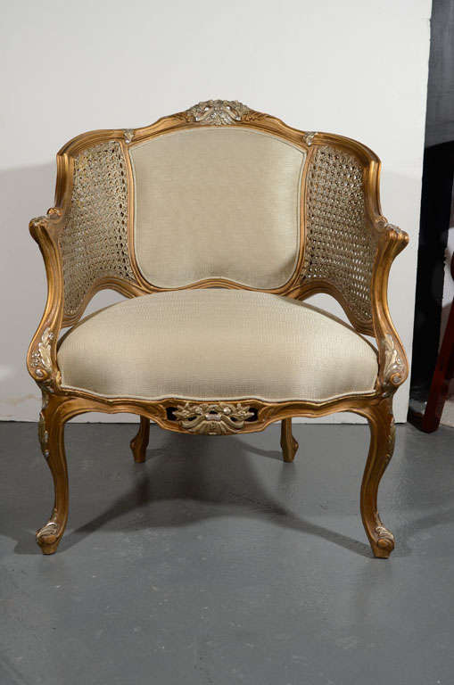 Hollywood Regency bergere chair in the style of Louis XV. Stunning hand-carved wood frame with gilt finish and details in white gold leaf. Chair has beautiful wide curvature form and features cane sides, and cabriole legs. Upholstered in woven