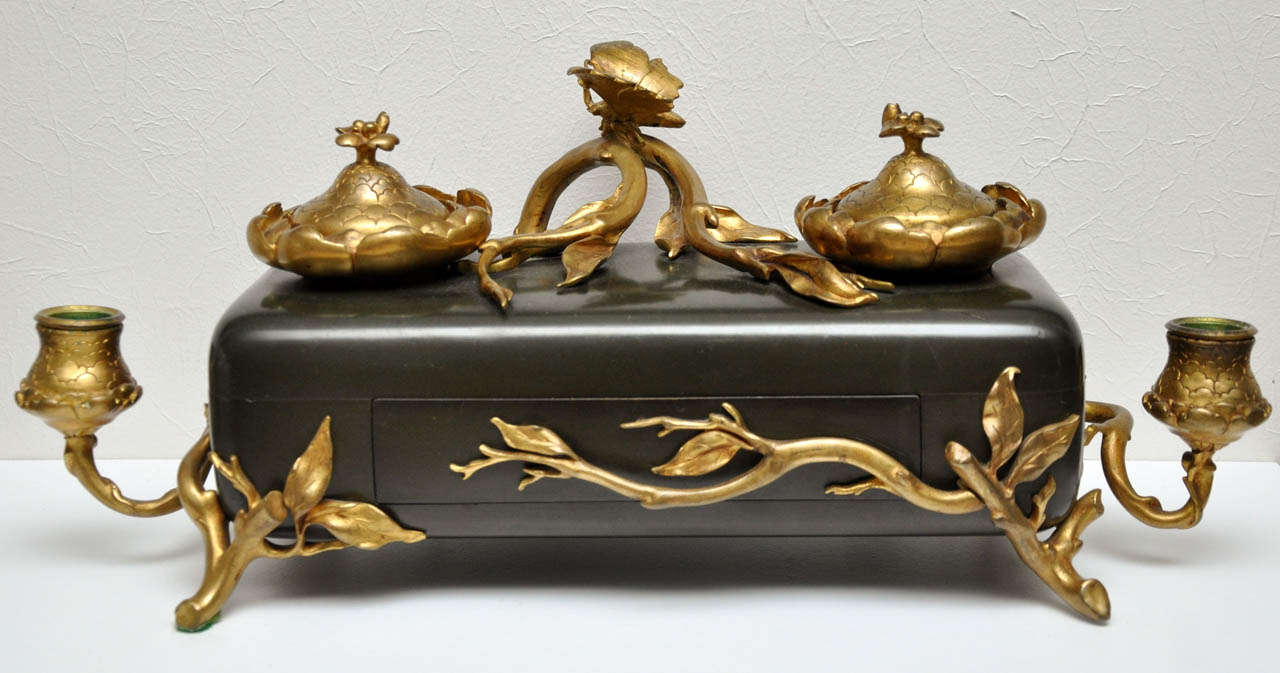 Brass flowers, vines, and a butterfly embellish the patinated base of this stunning Art Nouveau inkwell. Two brass candle holders flank a rectangular base with a small drawer.