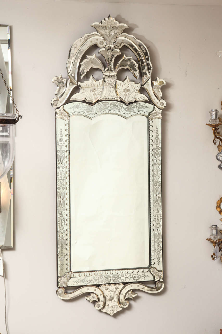 A vertical rectangular Venetian mirror, the frame having scalloped edged top surmounted by crest formed by leaf and scroll motifs. The mirrored frame in sections with fine detailed etched floral and leaf motif and having bulls' eye dots carved on