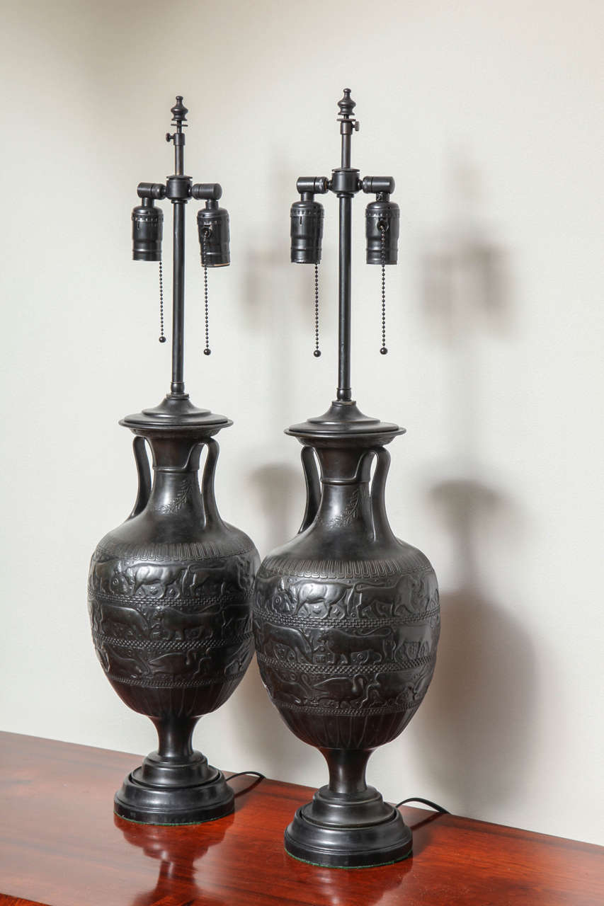 A pair of 19th century Grand Tour cast bronze table lamps. The bases in the form of an Assyrian inspired vessel with handles. The narrow waisted top and bulbous center tapering to a fitted wooden base. The body with low relief cast friezes of lions,