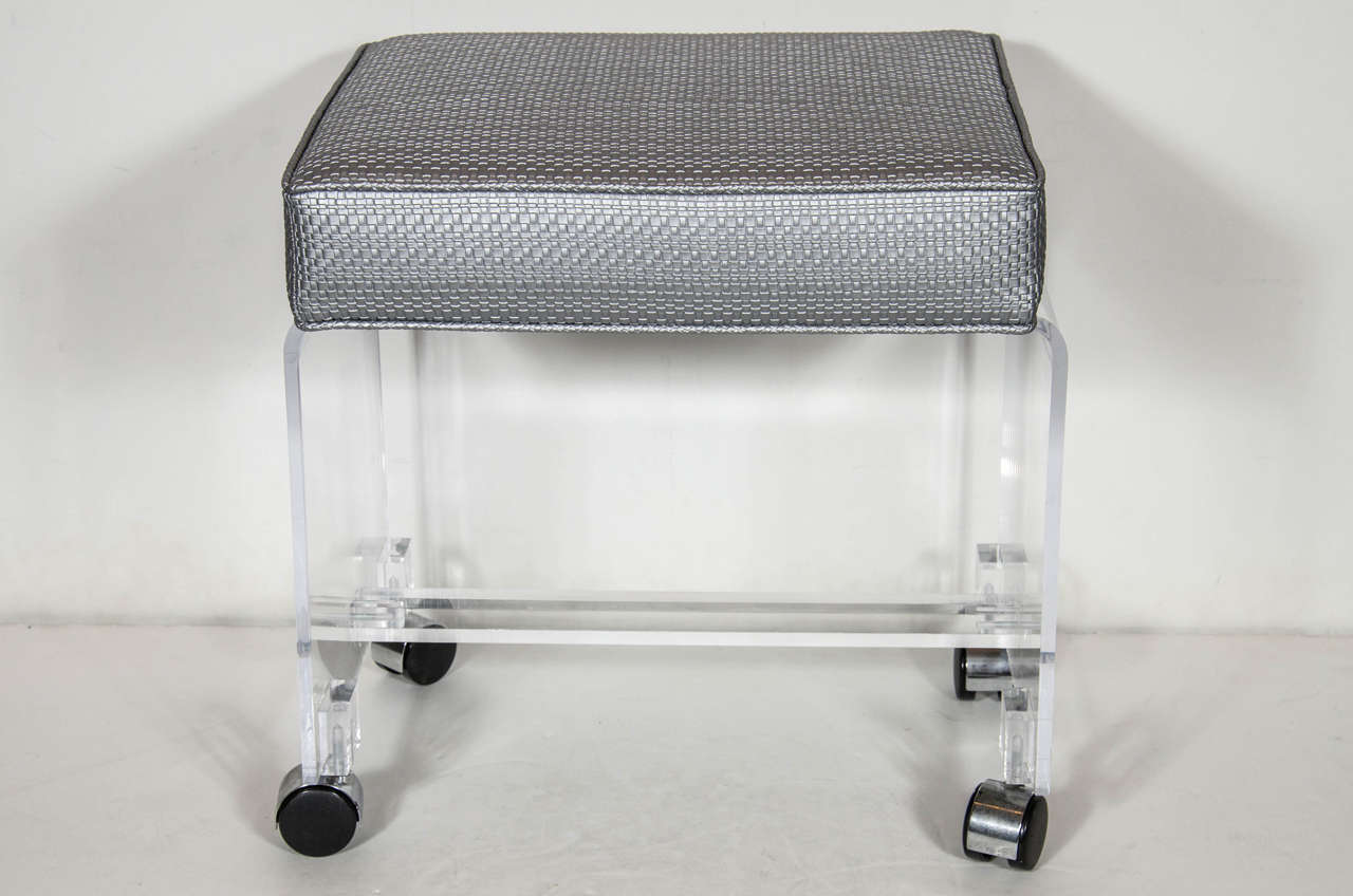 Chic Mid-Century Modernist clear Lucite bench or vanity stool in a waterfall design on chrome castors with a spherical cross support rod. The thick squared top cushion has been newly upholstered in a platinum metallic woven upholstery which makes