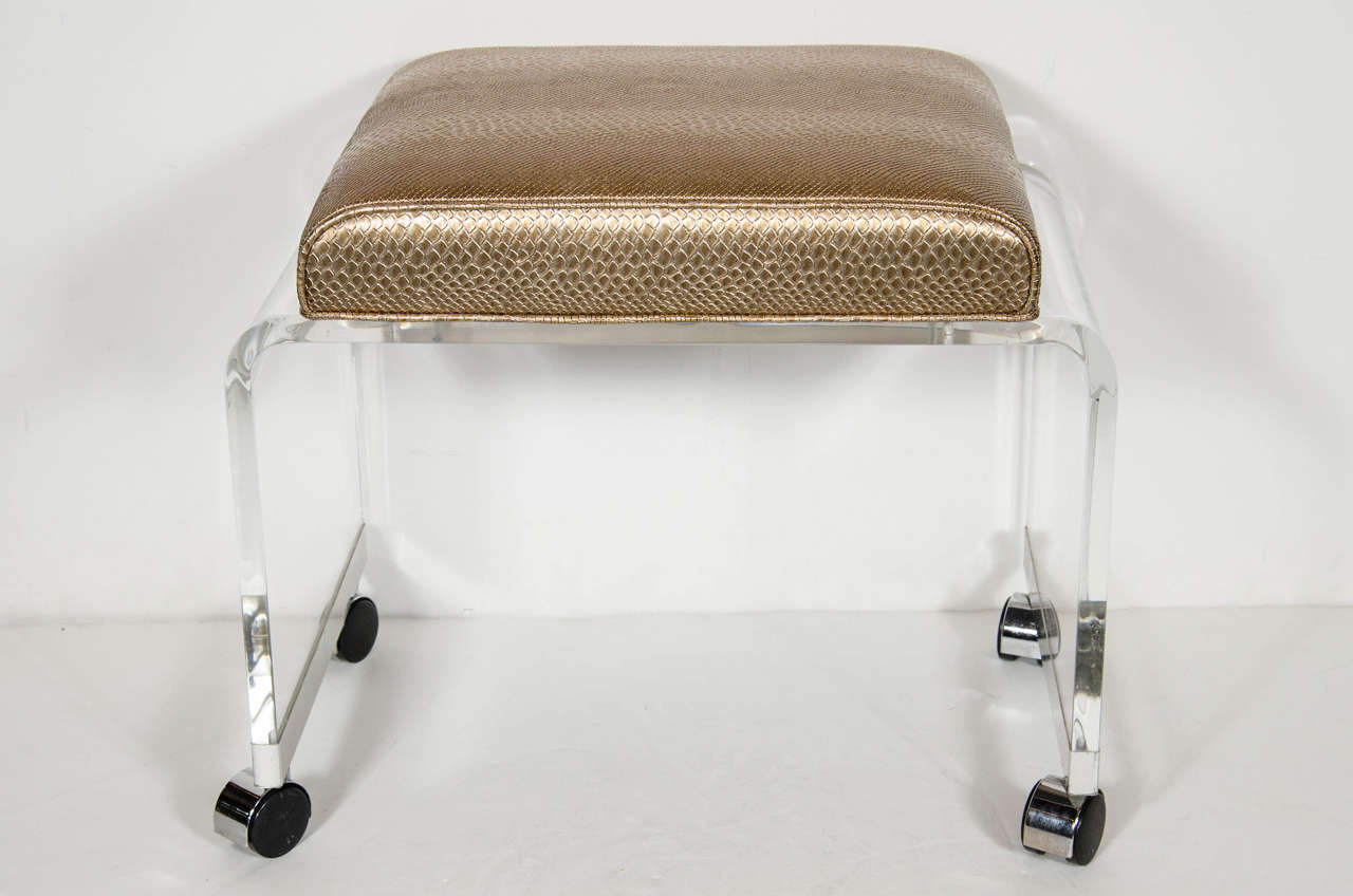 Chic Mid-Century Modernist clear lucite bench or vanity stool in a waterfall design with chrome banding on the base that support chrome castors. The top cushion has been newly upholstered in a faux bronze metallic python which makes this piece a