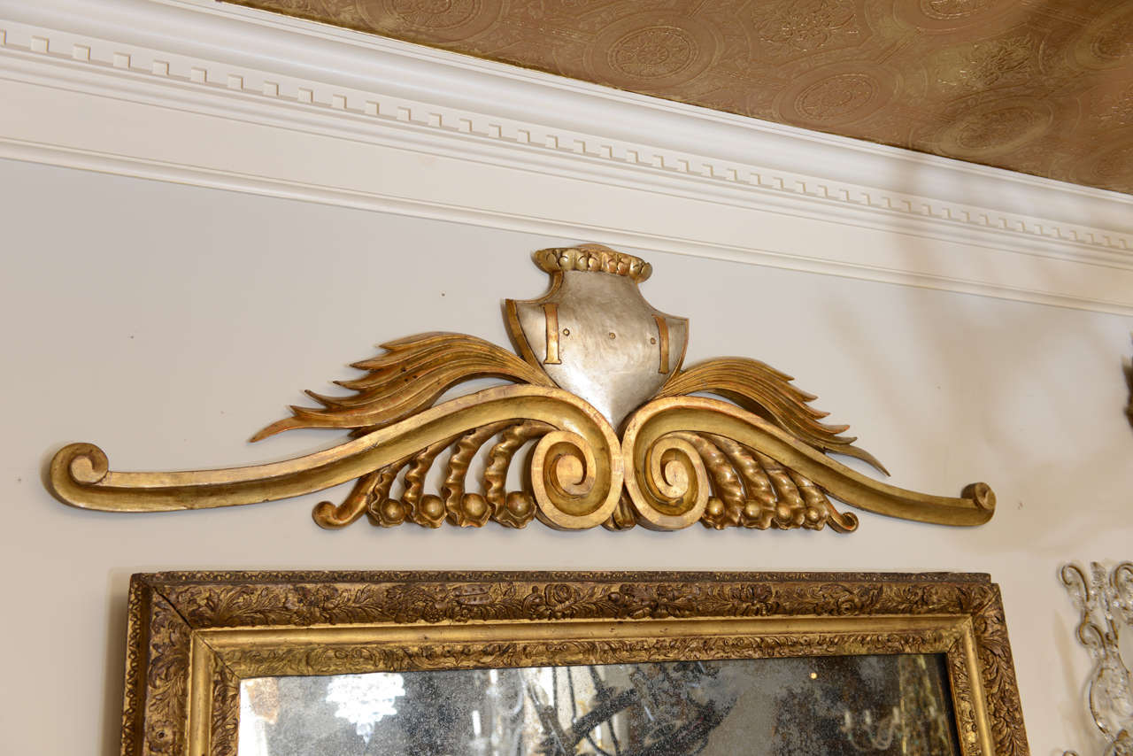Overdoor, of carved wood, having silvergilt shield surmounting a pair of elongated gold scrolls with leaf details.
