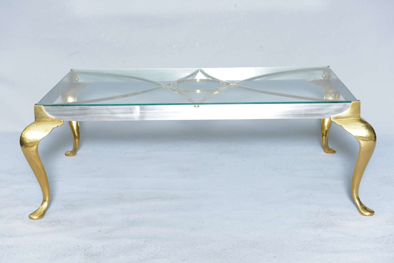 Cocktail table having a rectangular top of glass, on apron of polished steel, inset by decorative fretwork of brass, raised on stylized cabriole legs with pad feet. Rectangular top measures 42.25 in. x 26.5 in.