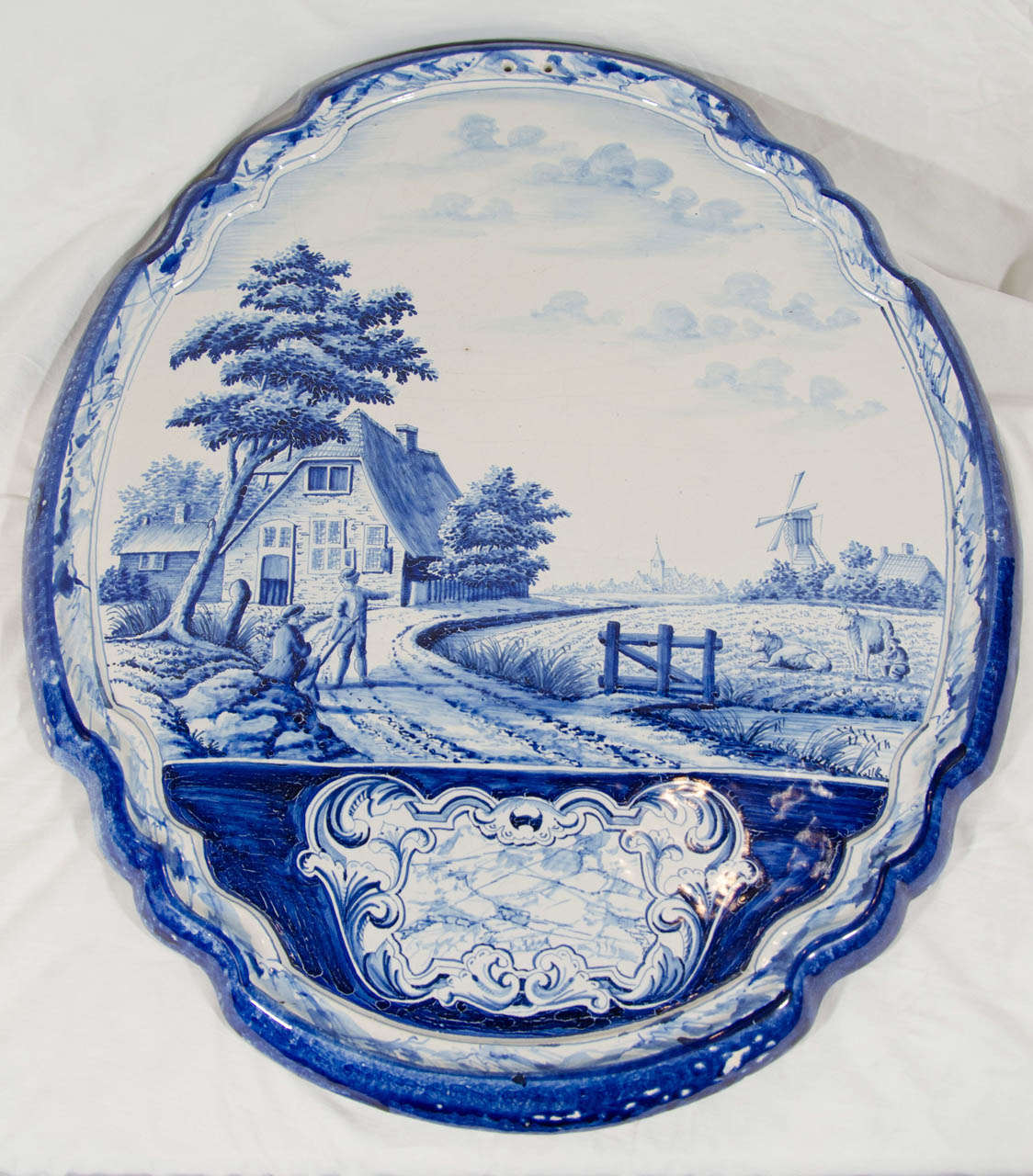 A large oval plaque painted in cobalt blue with a narrative scene of 2 gentlemen walking roadside, a cottage, a fence, and a field with cows. 
The plaque is pierced with two suspension holes for hanging.