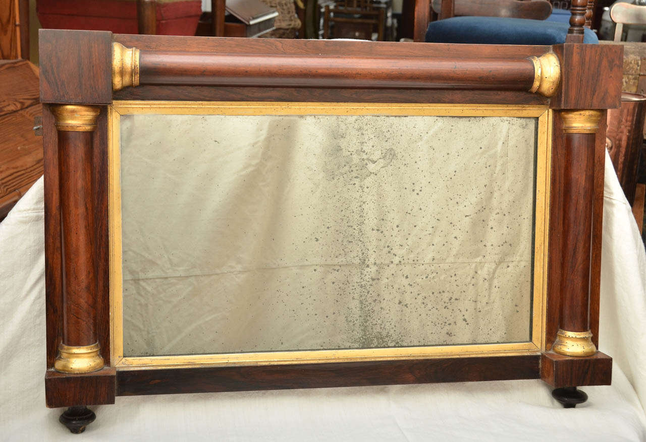 English Regency rosewood overmantel mirror with gilt capitols & gilt interior moulding surrounding the original mirror.  The mirror sits on 2 ebonized circular turned supports.  Pleasing distressing to the mirrors silvering.  Great over a tall chest