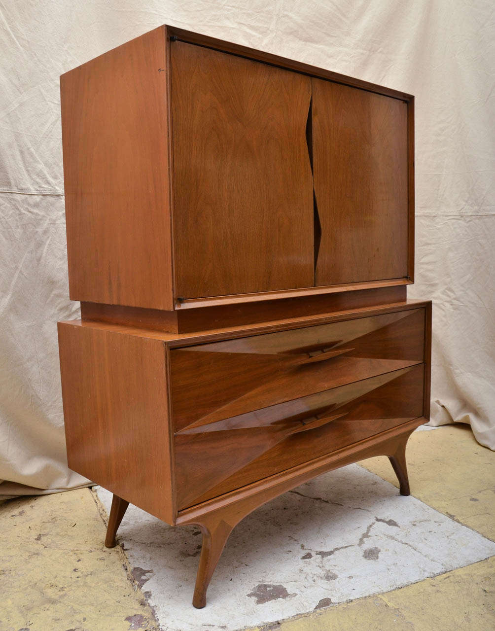 1960 Mid Century walnut two door cabinet with zig zag cut out that acts as door pulls.  Sitting on two drawer chest whose drawer fronts form an inverted pyramid design, centered by a narrow wooden pull. The chest is raised on four splayed legs. The