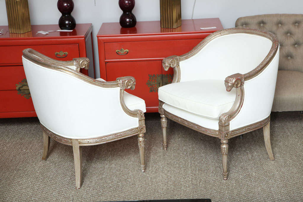 A pair of silvered wood and upholstered arm chairs with an elegant silhouette.  The chairs have beautiful carving on the curved back and seat rails, straight, turned front legs and splayed back legs, with rams' heads on arm rests.