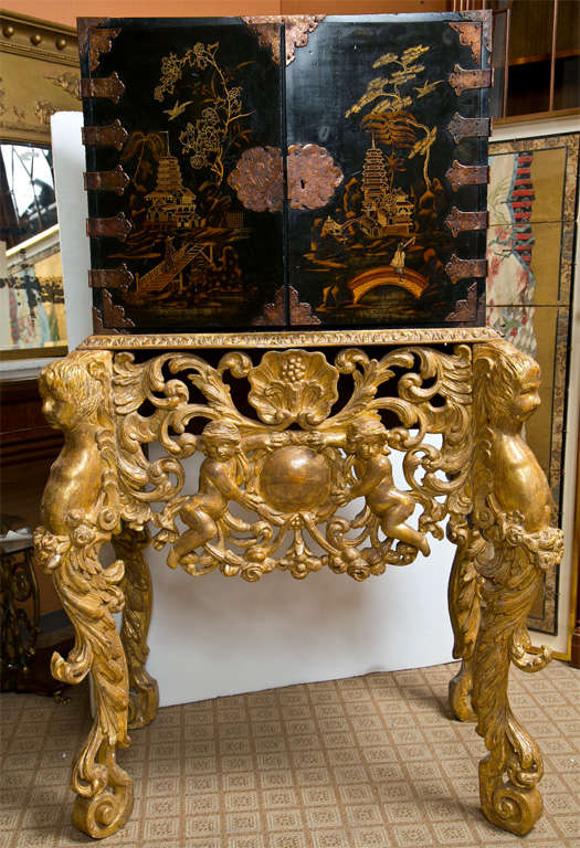 An intriguing 19th century Chinese treasure chest on a gilded base, the chest ebonized with hand painted detail, opens to reveal 6 small drawers and 2 small doors, raised on an intricately-carved gilded base decorated with cherubs, scrolls, and