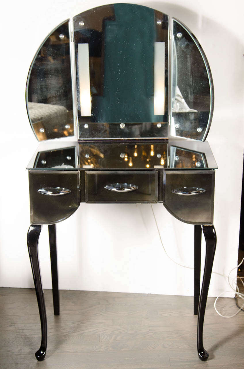 This stunning vanity features a stylish cabriole style leg of ebonized walnut .Vanity features three drawers with all smoked mirror and chrome pulls. The vanity also features a tri -fold illuminating mirror with hand beveled detailing.