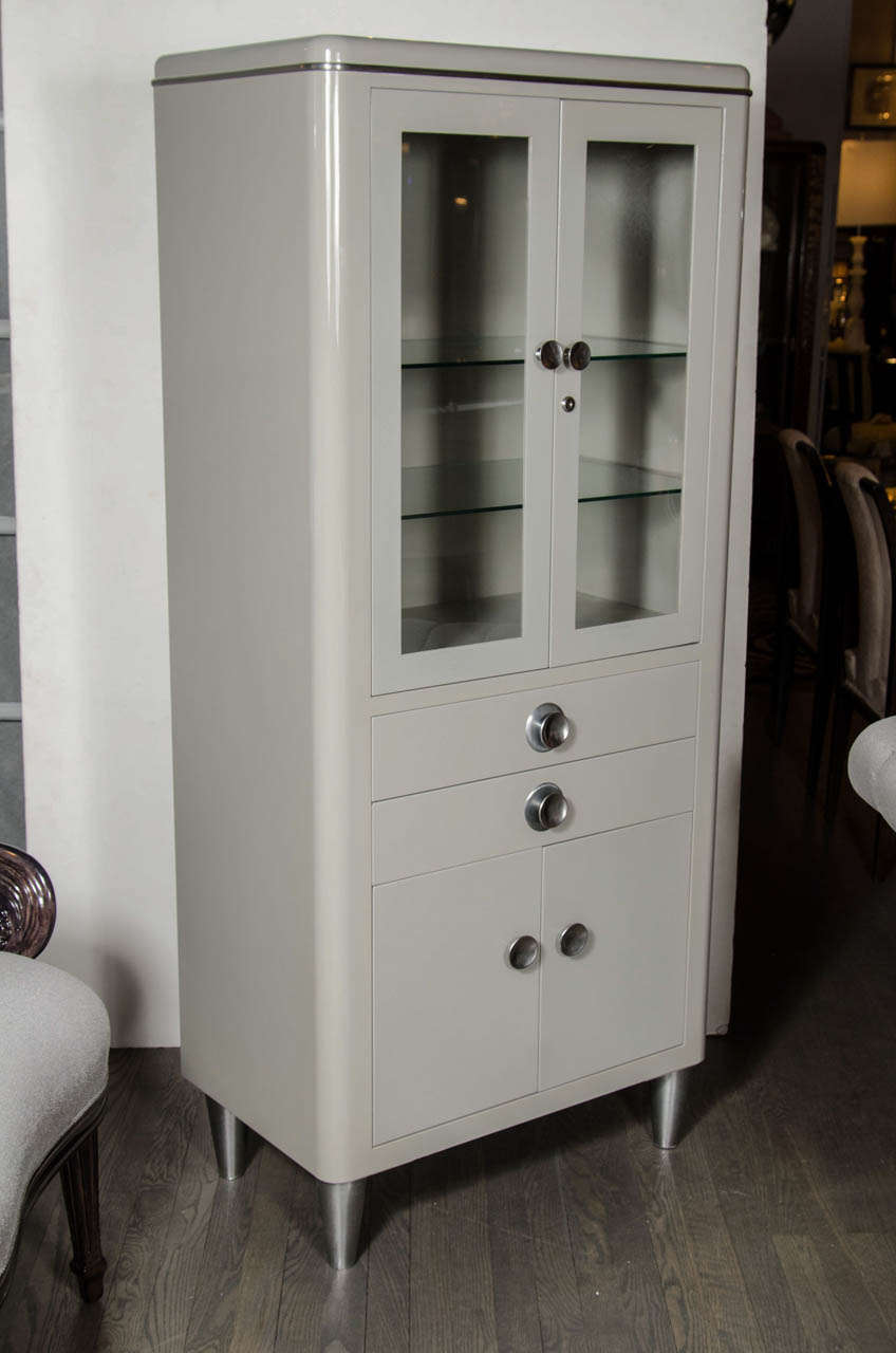 This Art Deco cabinet features two adjustable shelves and the interior illuminates as well. This cabinet is grey lacquered with brushed aluminum Banding,fittings and conical legs.It also has a lock and key mechanism on there doors.It also has two