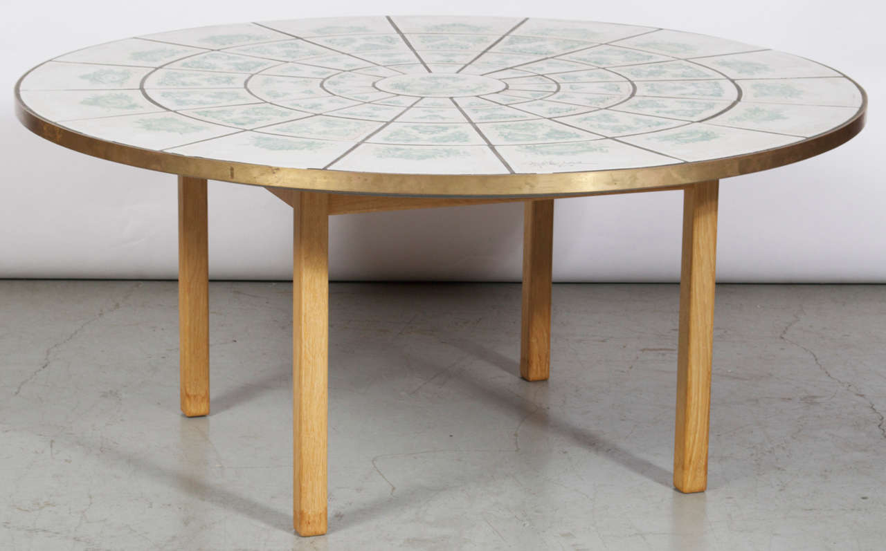 A coffee or center table by Bjørn Wiinblad. Four oak legs and a round, hand painted tile top. Tiles are rimmed by a brass band. Signed and Dated 1970.