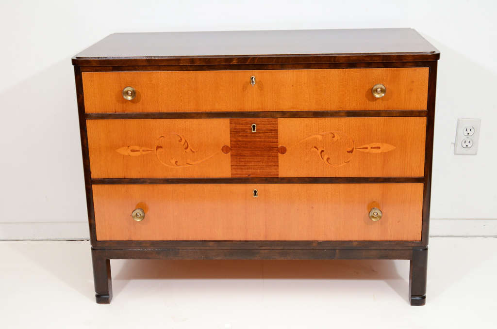 A compact and sturdy locking three drawer chest, with walnut-stained birch case, contrasted with golden-ash veneered drawer faces. The center drawer is adorned with a central palisander panel and balls, from which swirling stylized vines and leaves