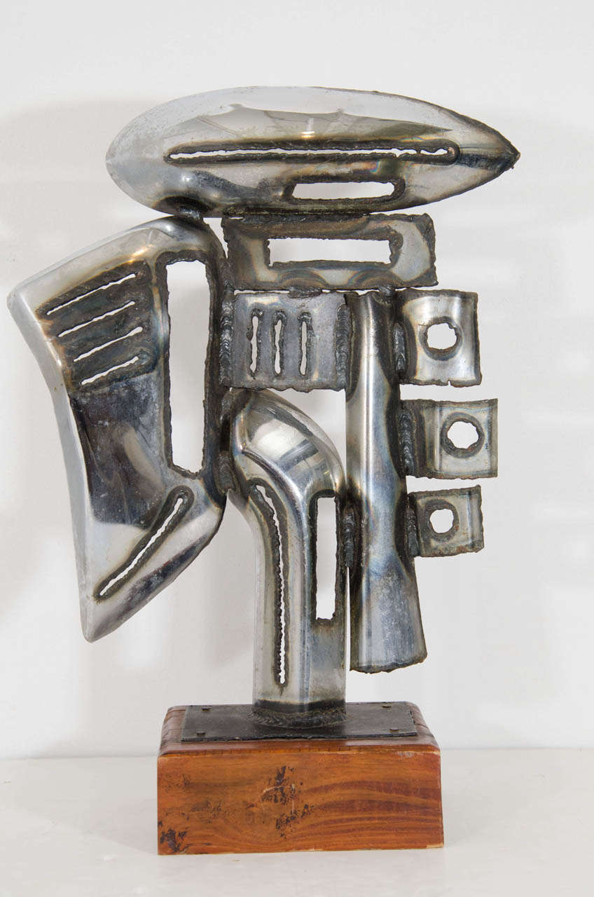 A vintage sculpture made of chrome car parts and mounted on a wooden cube base.  Illegibly signed. The signature appears to have the first initial "T" and the last initial "P."

Good vintage condition with age appropriate wear.