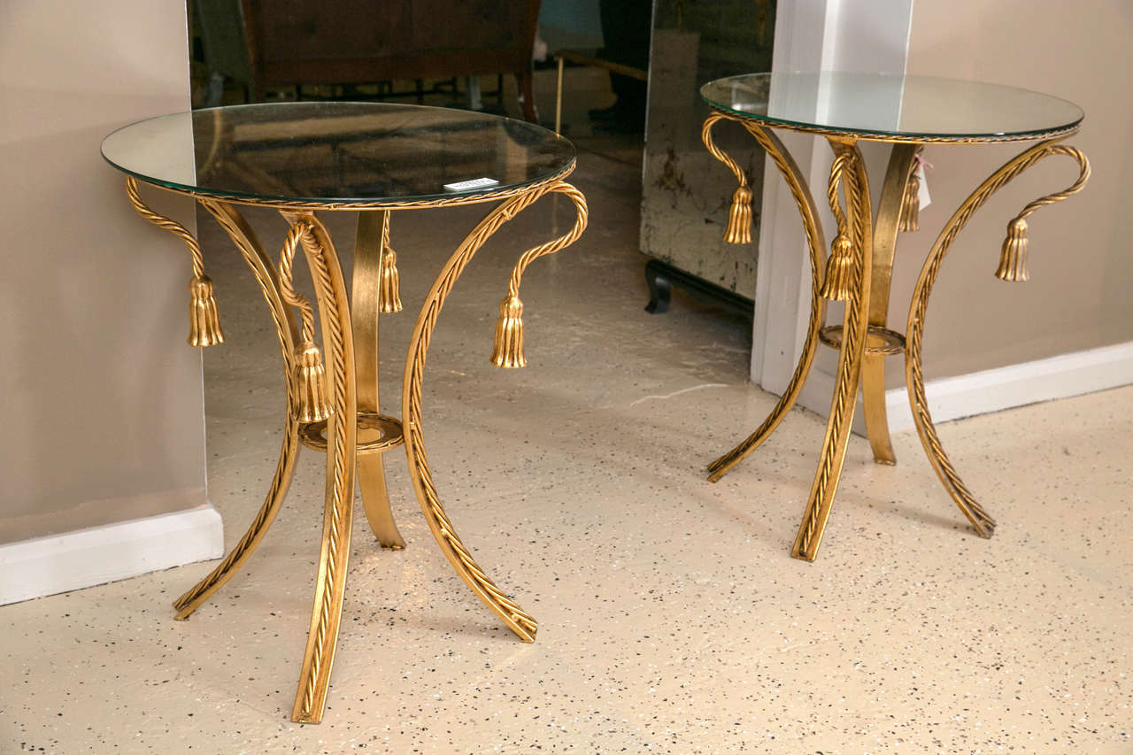 A fine pair of tassel form and rope edge mirror top end tables. The curved brass rope form legs leading to a brass circular top with four tassel ornaments supporting a mirror top.