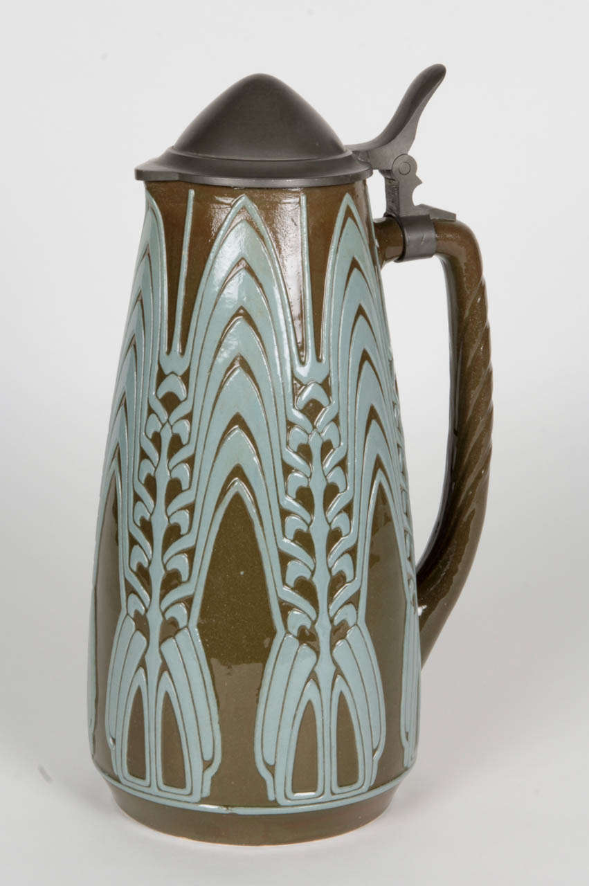 PETER BEHRENS  (1868-1940)  Germany
SIMON PETER GERZ (founded 1862) Höhr, Germany 

Henkelkrug / beer pitcher  c. 1907

Salt-glazed stoneware pitcher with blue-grey stylized graphic motif on a 
loden green body, mounted pewter lid 

Marks: