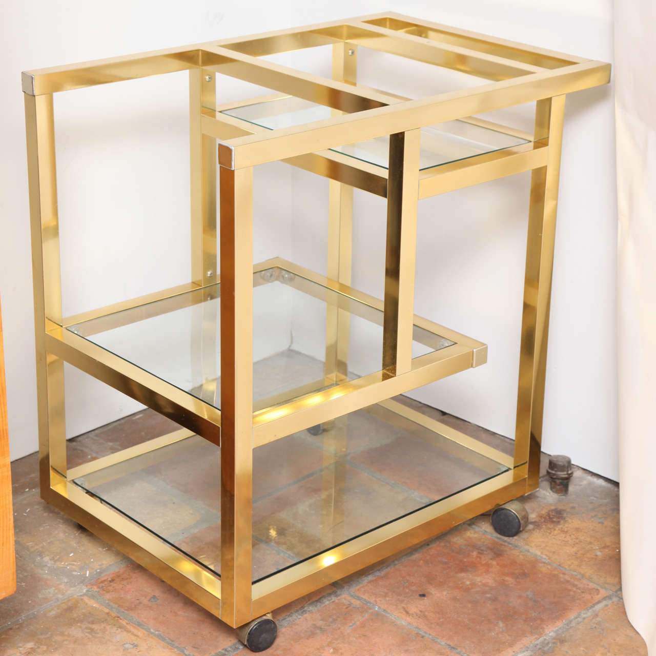 A stylish vintage modernist bar cart in brass with three lightly smoked glass shelved tiers in a practical cut-out configuration. The cart sits on easy rolling sealed covered casters. Designer is unknown in the style of Milo Baughman.