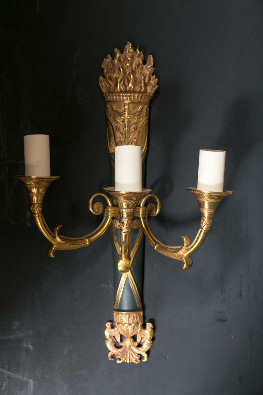 Each sconce wired. Three trumpet form arms hold the lights. The back is of patinated bronze with criss cross design. The top with a flame form above a capital. The bottom finial of acanthus leaves and griffins. May be sold as pairs.