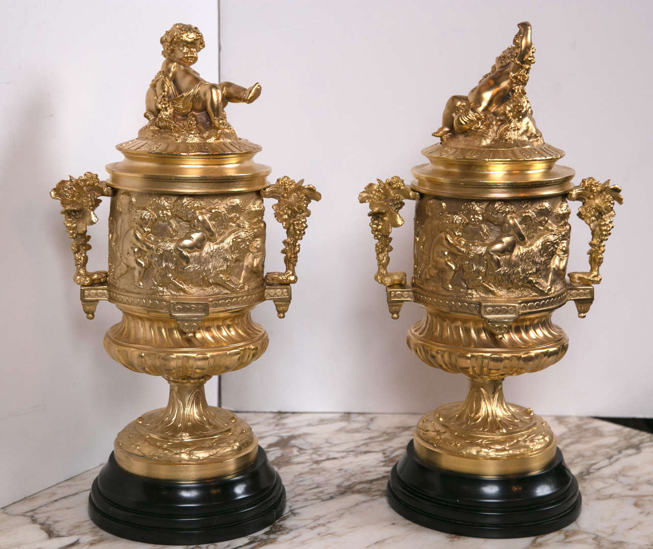 Raised on turned black stone bases, this pair are decorated in the Clodion manner with putti and animals.  Each urn has a lift out liner. Each lid is surmounted with a putto. The arms  designed with grape leaves and grape clusters.