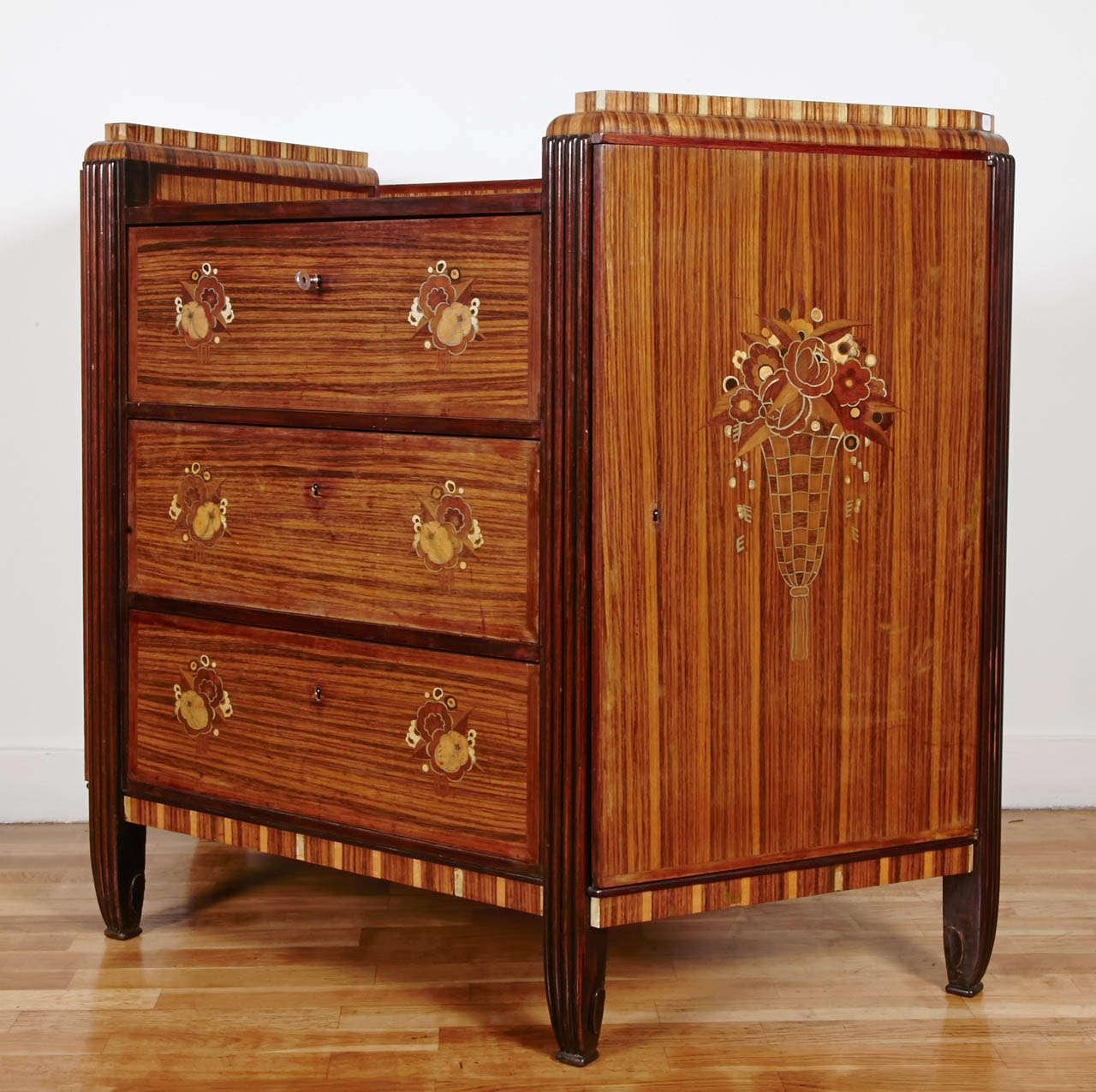 1925's rosewood veneer commode with three front drawers adorned with stylized floral motives in exotic woods and metal. On both sides of the central body the two doors are adorned with center motive of a vase of plenty. A rosewood and ivorine spine