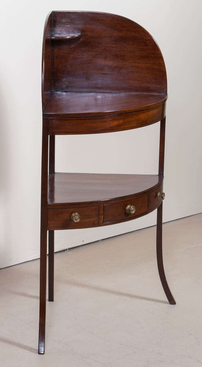 A gorgeous, Regency mahogany corner wash stand. This bow-front washstand has a splash back with a tiny top shelf. The center open shelf has a single frieze drawer flanked by two dummy drawers. The stand ends on square supports with splayed legs.