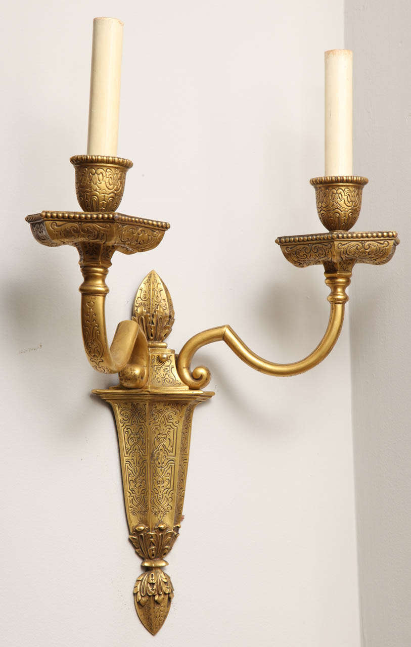 A pair of Caldwell two light Dutch Baroque style gilt bronze sconces with incised decoration over all. Each candle arm with shaped bobeche having incised decoration and finial at the bottom of each tapering backplate.